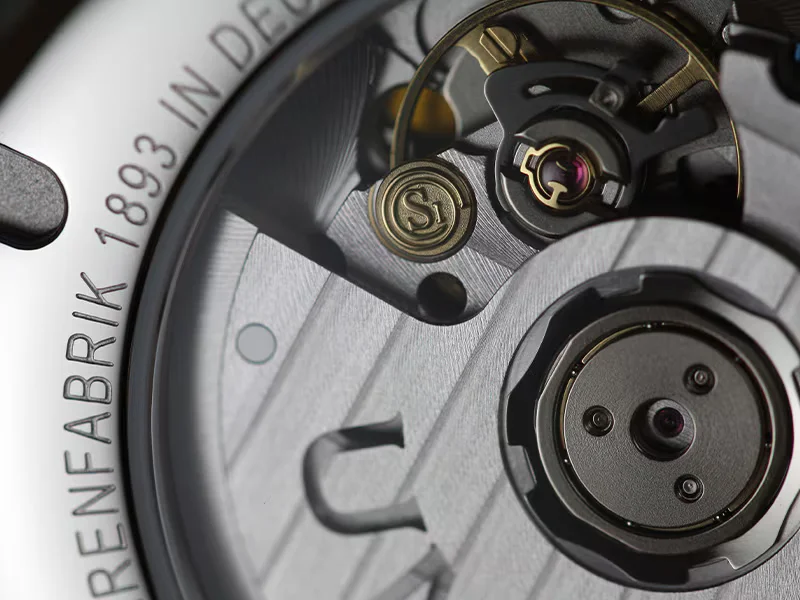 Macro shot of a watch movement with Geneva stripes, ruby jewel, gold S1 screw, and "FABRIK 1893" engraving.