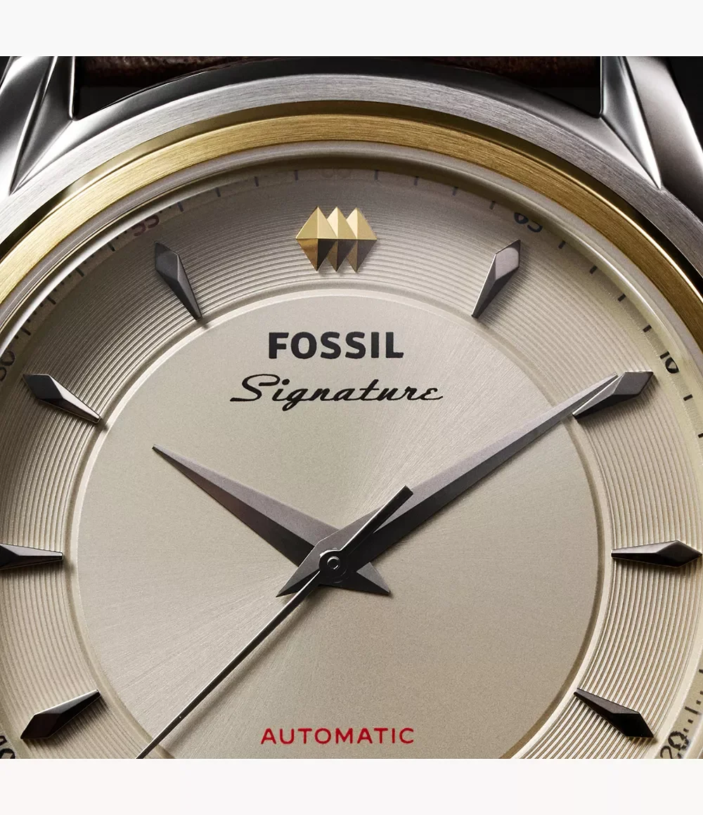 Close-up of Fossil Signature automatic watch. Textured silver dial, gold bezel, dark markers, gold emblem, brown strap.