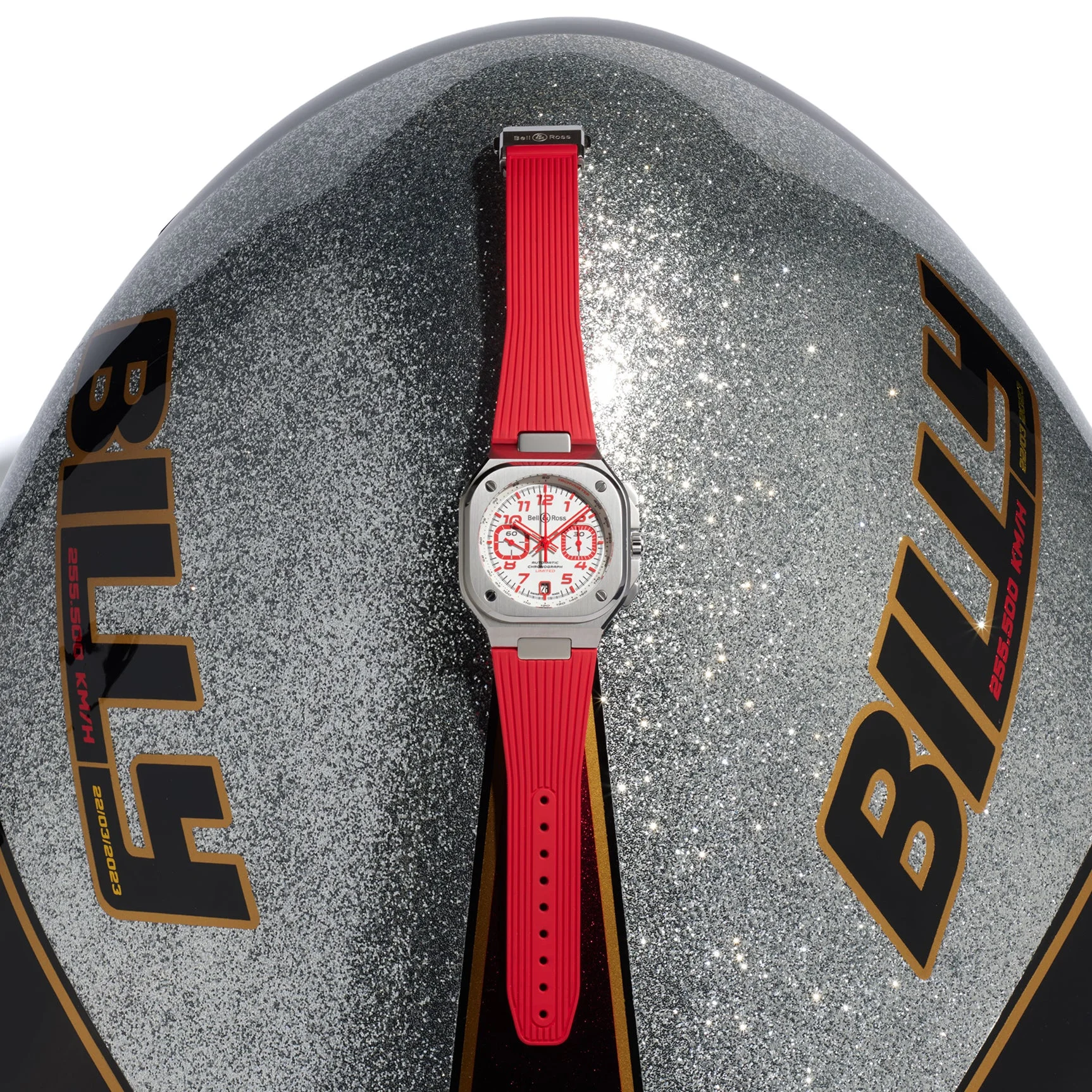 Square Bell & Ross BR 05 Chrono with white dial, red accents, and red rubber strap on a glittery "BILLY" racing helmet.