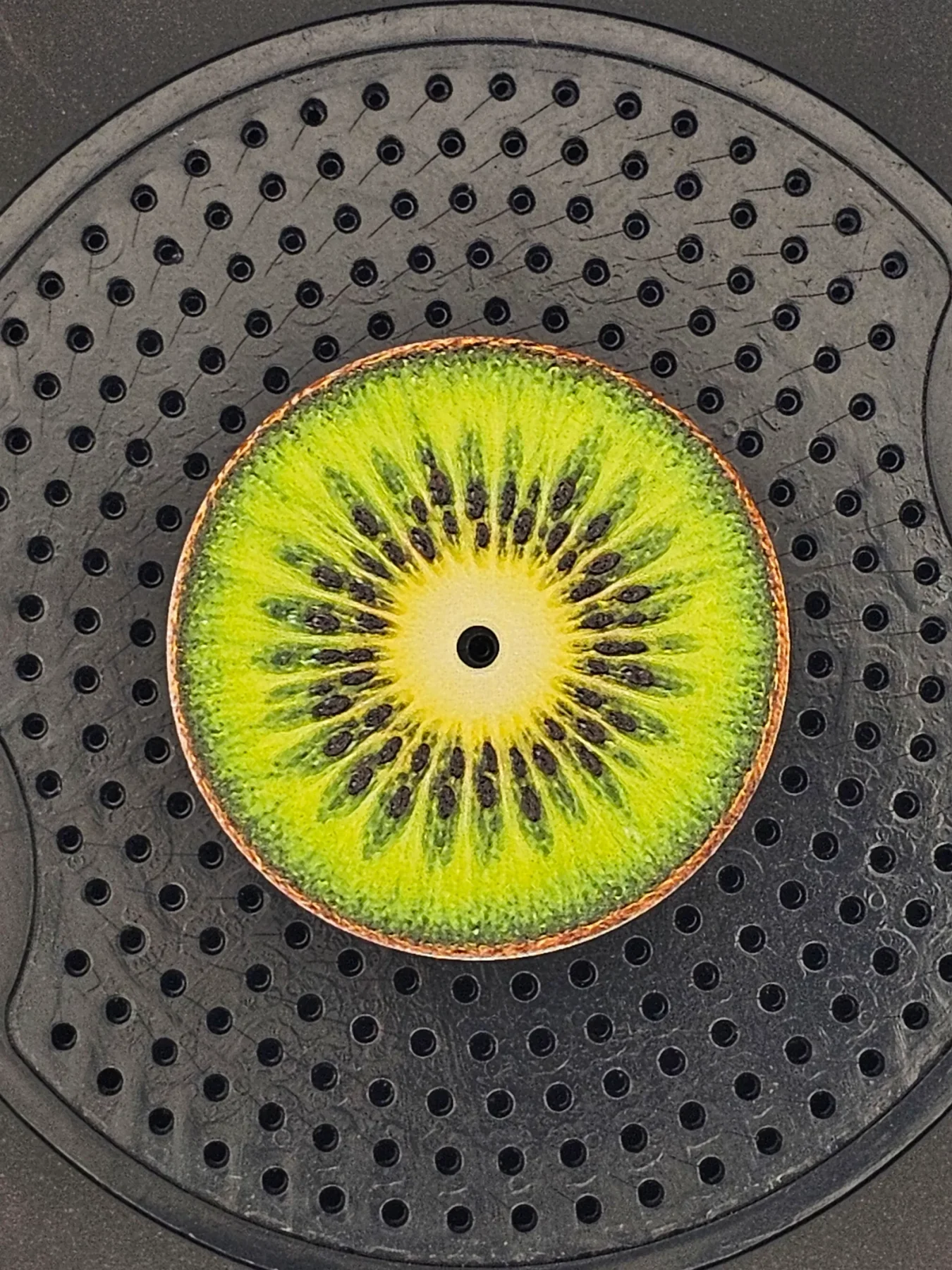 Vibrant green kiwi-slice watch dial with a central pivot hole, on a textured black grid surface.