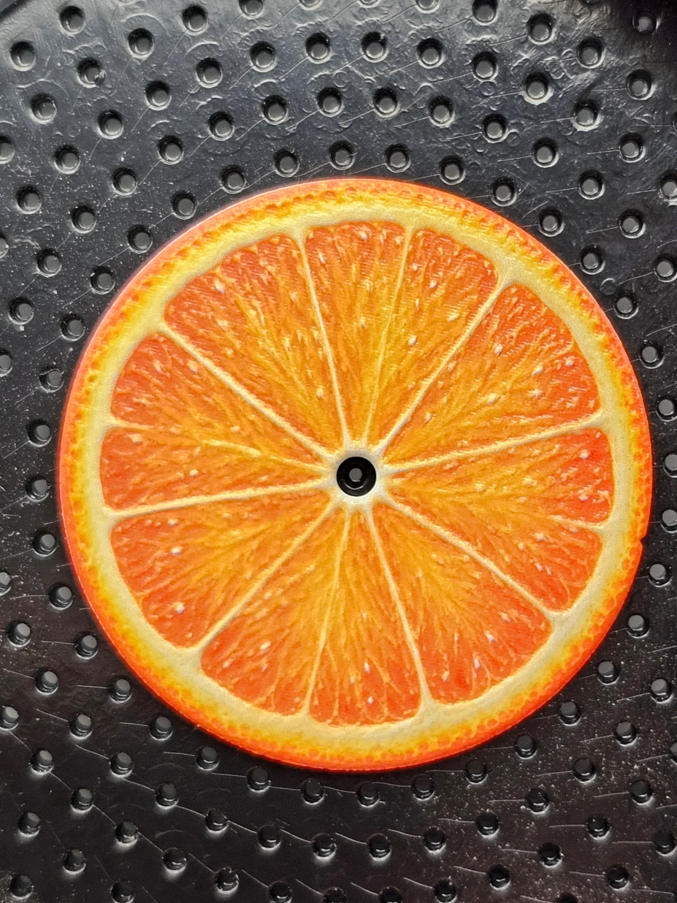 Circular item depicting an orange slice, with a black center hole, on a black perforated mat.