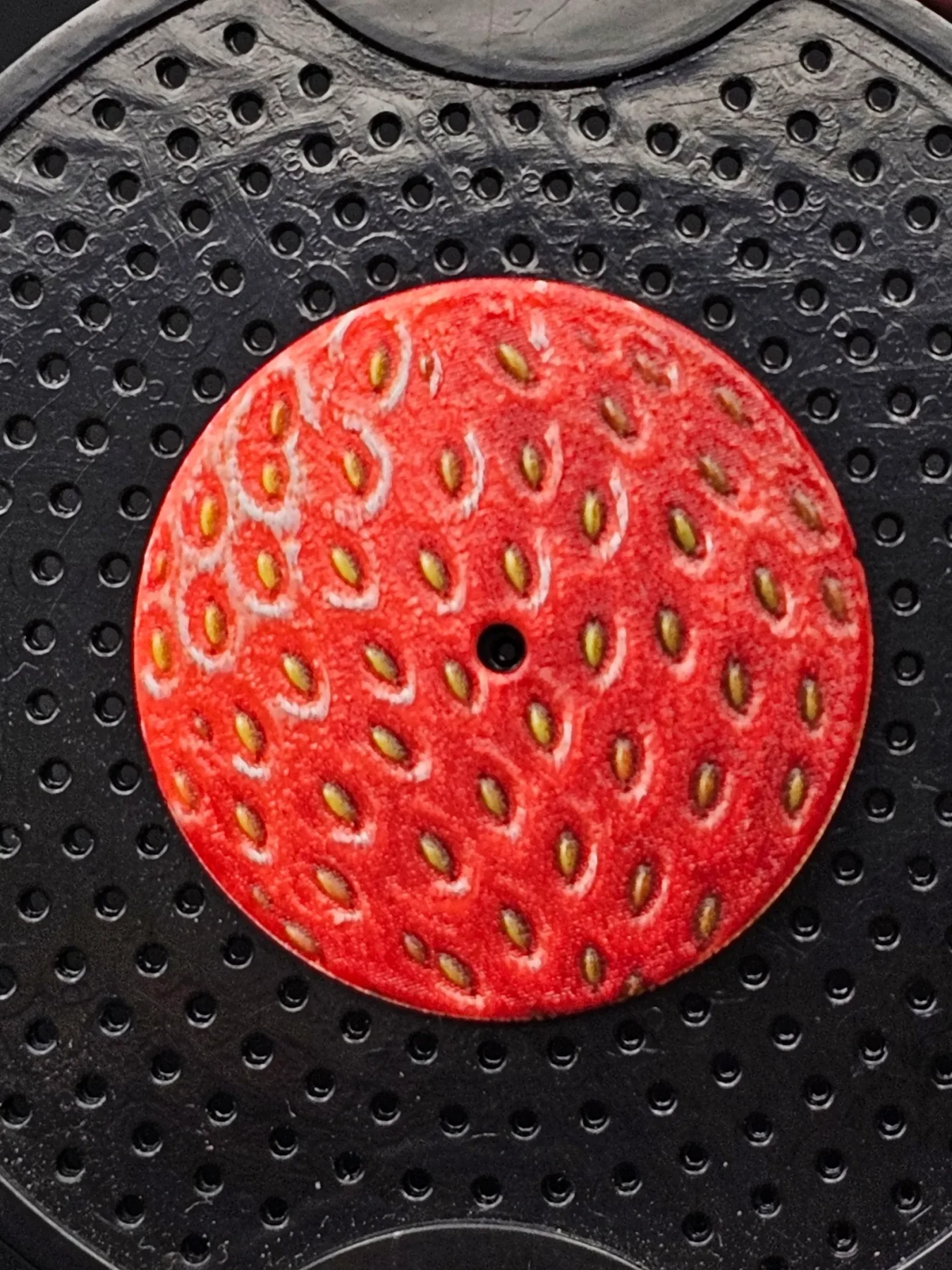 Round red watch dial with realistic strawberry texture and yellow seeds, on a black dimpled surface.