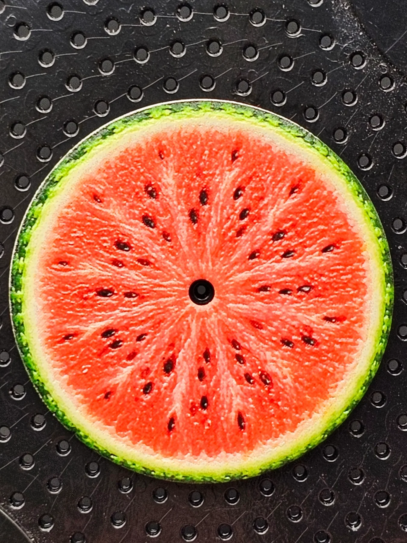 Circular watermelon motif with bright red interior, black seeds, green rind, and central black hole on textured black surface.