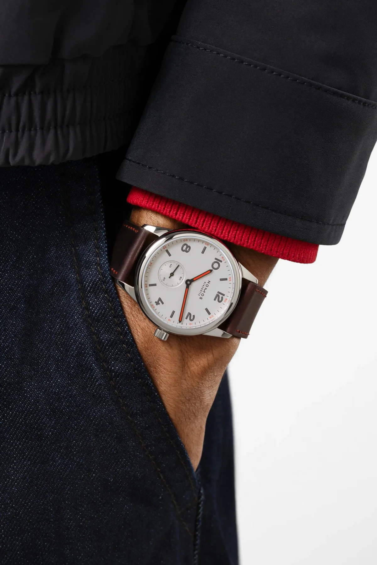 Hand in pocket shows Nomos Glashütte watch with white dial, red hands, brown leather strap. Dark jacket, red sweater, jeans.