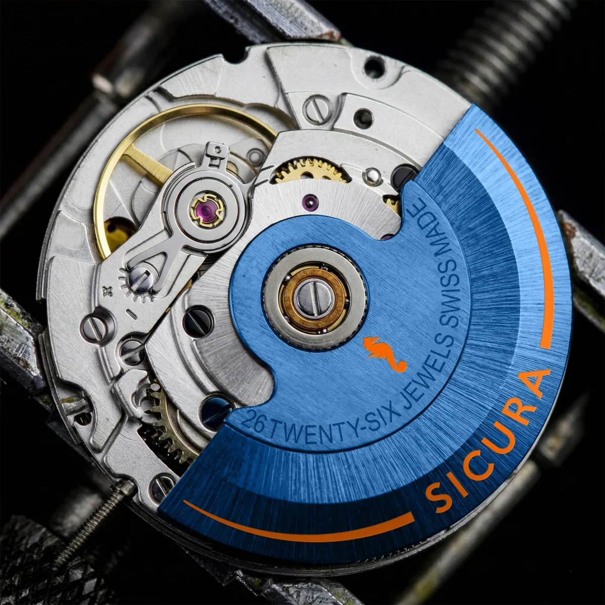 Macro shot of an automatic watch movement with a blue rotor, orange "SICURA" text, gold balance wheel, and jewels.