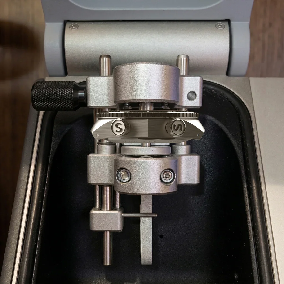 Watch component with twin 'S' logos held by a silver precision machine for testing or calibration.