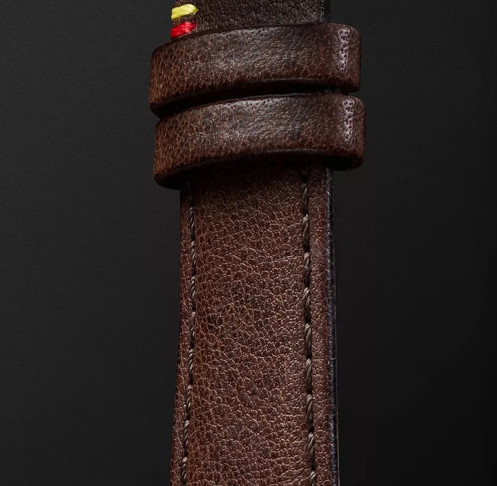 Close-up of a brown leather watch strap with two keepers & multicolored stitching details on a dark background.