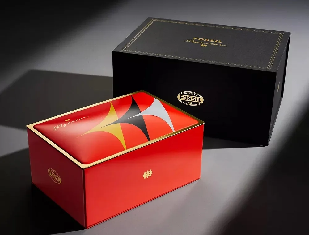Two Fossil Signature boxes on a dark surface. Black box with gold text, red box with gold trim & colorful starburst lid.