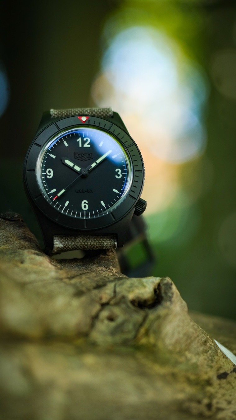 Monday battle wristed with a masterclass in rugged microbrand horology, striking a perfect balance between bold 1960s diver aesthetics and modern tactical resilience. Greg Stevens, widely celebrated within the watch community for crafting world-class custom leather watch straps since 2005, brought his “less is more” design philosophy straight into this heavy-duty, no-nonsense tool watch. For those seeking an affordable, grab-and-go mechanical timepiece that stands out from the sterile, mass-produced crowd, the PVD-coated 5 Series represents exceptional value and undeniable wrist presence.
The strap force is strong with this one!
You cannot talk about a GSD watch without highlighting the strap. Greg Stevens leveraged his two decades of leathercraft expertise to deliver an unparalleled unboxing experience. The 5 Series features a 22mm lug width, specifically designed to accommodate substantial, high-character footwear.
The PVD 5 Series is frequently paired with a vintage military canvas strap. Often cut from authentic, mid-century military equipment—like 1950s French ammo pouches—the canvas adds incredible texture, tells a historical story, and patinas beautifully the more it is exposed to the elements.
The Greg Stevens Design 5 Series PVD is the ultimate “workshop watch”—the piece you strap on when you want to get your hands dirty without second-guessing its durability. It competes toe-to-toe with established independent tool-watch brands like Resco, Nodus, and Raven, but wins out for enthusiasts who value hand-crafted, artisan straps and highly limited, bespoke execution.