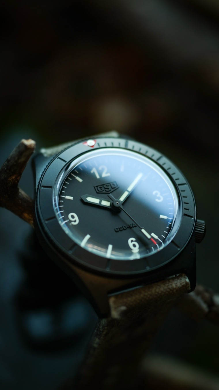 Going into Monday battle with the GSD 5 series. Originally known for creating world-class, custom-tailored leather straps, artisan Greg Stevens engineered this microbrand tool watch to serve as the ultimate high-durability “workshop piece”.

The entirely black PVD (Physical Vapor Deposition) coating transforms the 42mm architecture into a stealthy, light-absorbing tool. To maximize comfort during active use, the fluted screw-down crown is thoughtfully tucked away at the 4 o’clock position. This design choice prevents the crown from digging into the back of your hand when you bend your wrist.

The Ghost-Branded Dial
One of the most praised aspects of the 5 Series is its approach to minimalism. The matte black dial features a highly legible, classic layout. However, the real magic is in the branding: the GSD logo and text are subtly embossed directly into the black surface. Under direct light, the dial looks entirely sterile and tactical; angle it slightly, and the ghosted branding reveals itself with premium, reflective depth.

Low-Light Visibility
The hands and hour markers are generously layered with Superluminova (available in standard C3 or a vintage-toned “Old Radium” finish), offering immediate legibility in zero-light environments.

You cannot fully appreciate a GSD watch without highlighting the footwear. Because Greg Stevens is a master strap maker, buying a 5 Series includes a custom, premium strap of your choice.

While users can opt for premium Horween Dublin or English Bridle leathers, the PVD variant looks exceptionally striking when paired with his signature vintage military canvas. Often cut from authentic, mid-century military gear (such as 1950s French or Swiss ammo pouches), these straps offer incredible texture, tell a historic story, and develop a beautiful, rugged patina over time.
.