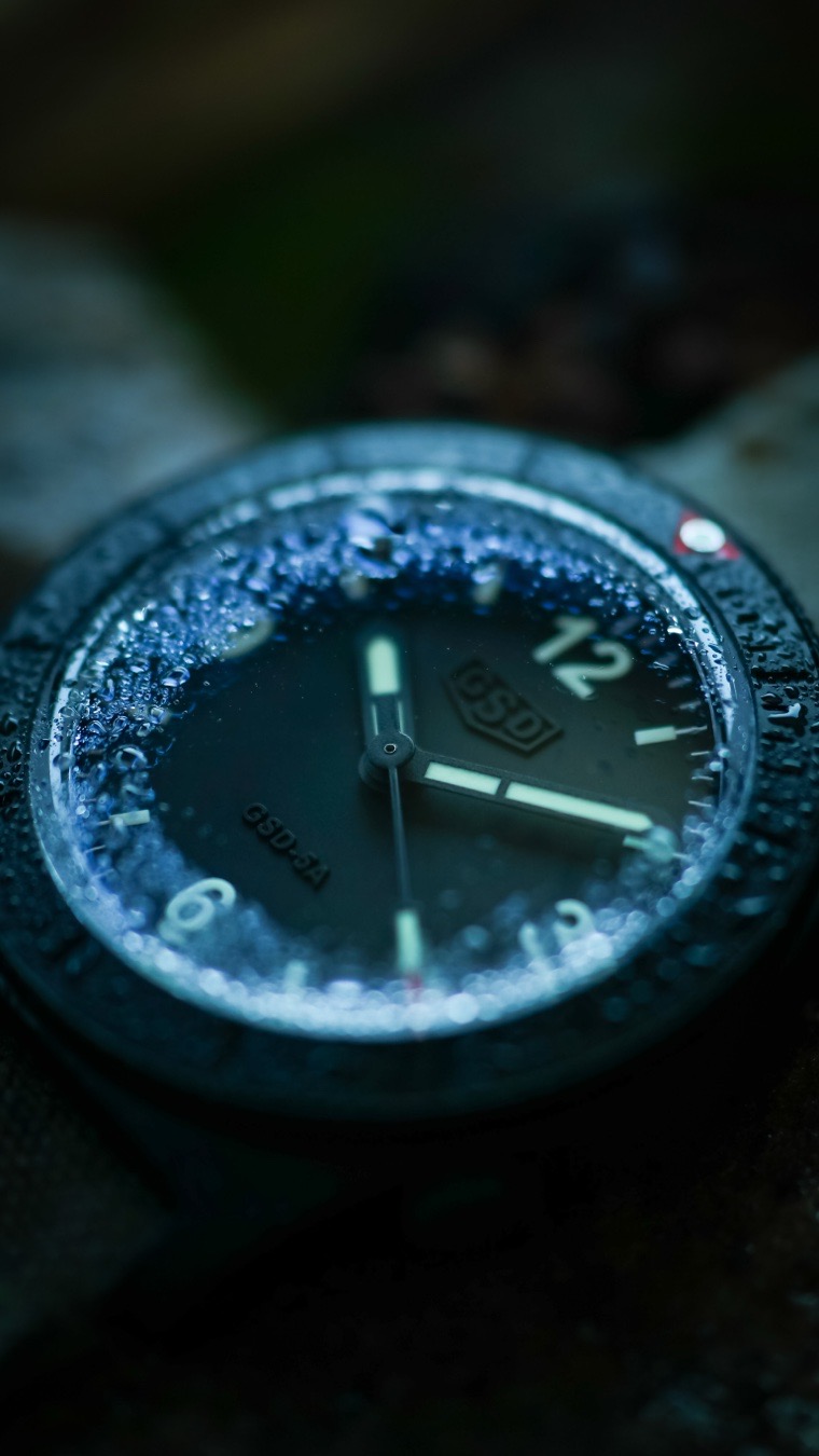 The Greg Stevens Design (GSD) 5 Series successfully bridges two distinct horological worlds, functioning as a true hybrid field watch diver. While it is officially categorized as a diver due to its robust architecture, its design language, minimalism, and pairing choices heavily channel the spirit of a classic military field watch.
Here is a breakdown of how the GSD 5 Series seamlessly blends these two identities into the ultimate “workshop watch”.

Diver Architecture, Field Watch Aesthetic
At its core, the 5 Series provides the uncompromising utility of a heavy-duty dive watch. It features a 42mm case, a beautifully domed sapphire crystal, a screw-down crown, and a massive 300 meters of water resistance (30 ATM). A reliable Swiss Sellita SW200-1 automatic movement powers the watch.

However, the visual execution skips the loud, flashy polished elements of a traditional luxury diver, pivoting instead toward the utilitarian, non-reflective nature of a field watch. 

It achieves this through specific finishes:

*The dial features a stark, high-contrast, sterile layout. The GSD logo and text are subtly embossed directly into the matte black surface. This “ghost branding” prevents any distracting reflections, mimicking a tactical field instrument.

*Available in stealthy PVD or a specialized “weathered” finish, the case has the rugged, matte appearance of a piece of land-based military gear.

*The hands and markers use thick layers of C3 or “Old Radium” Superluminova. This delivers the instant, low-light readability required for both midnight diving and night-time field operations.

A standard field watch is loved because you can get it dirty without second-guessing its survival. By packing 300M dive specs into a matte, ultra-legible, military-strapped package, the GSD 5 Series removes the vulnerabilities of both genres. It won’t flood if you jump in the water like a standard field watch might, and it won’t scratch or flash in the sun like a desktop diver. It is a dual-purpose watch meant to be beaten up.
.