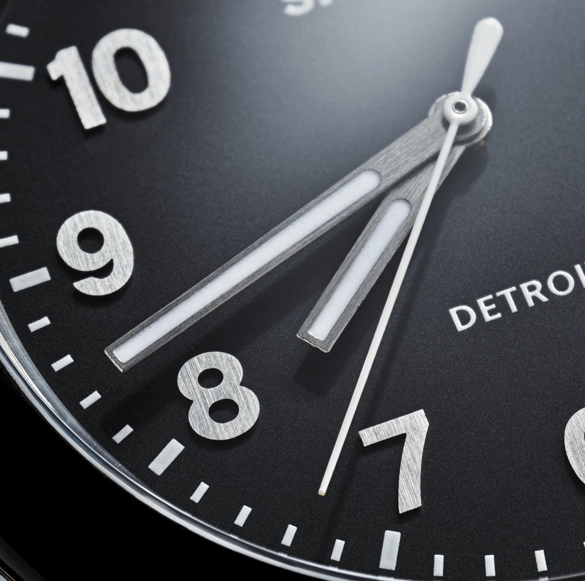 Close-up: black Shinola Detrola watch dial. Brushed silver numerals 7-10, luminous hands, white second hand. "DETROIT" branding.