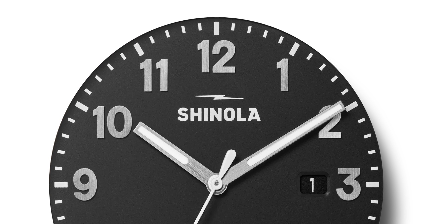 Close-up of Shinola watch. Matte black dial, bold silver Arabic numerals, white minute markers, silver hands, date '1'.