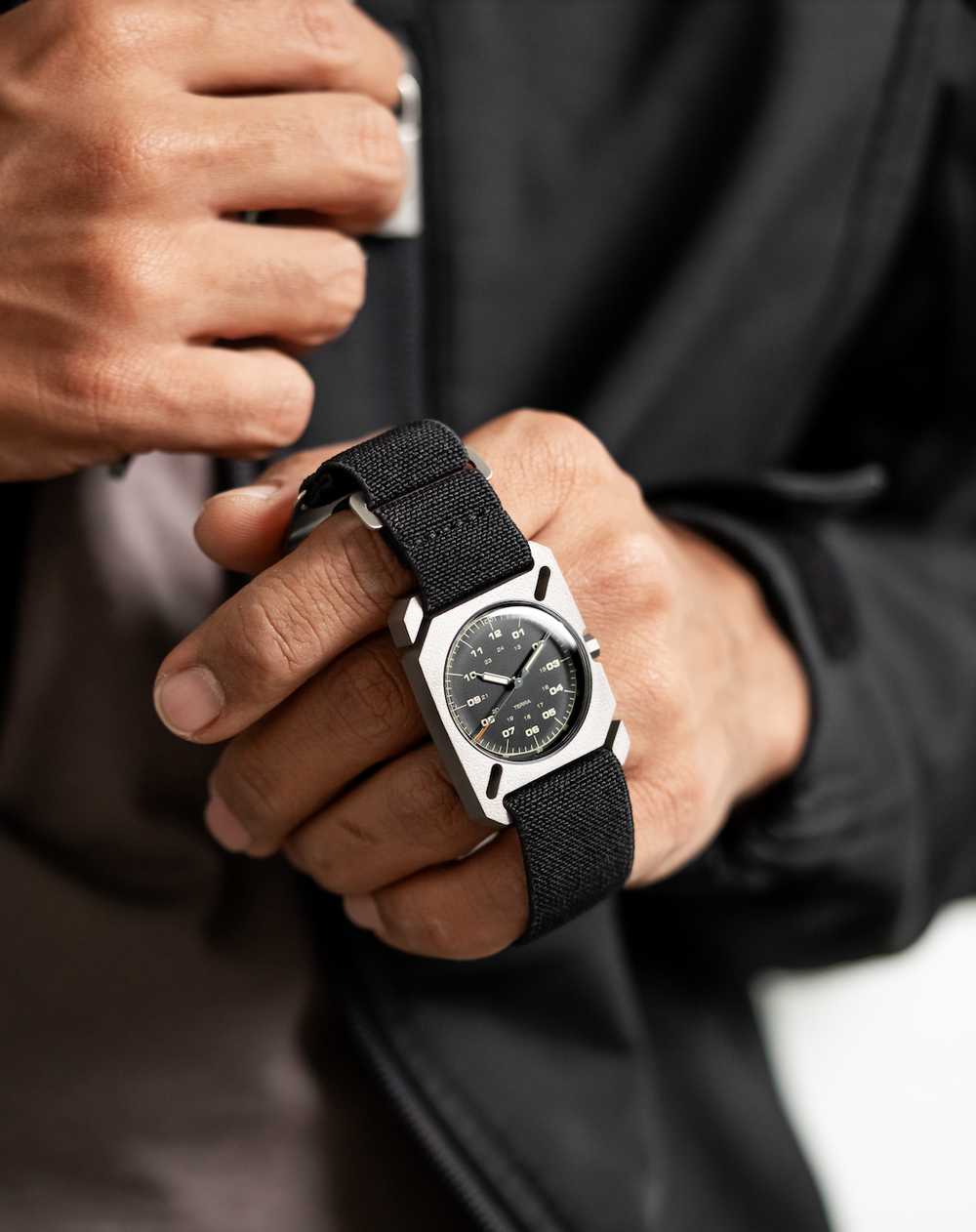 Hands hold DWC Terra watch: raw square titanium case, dark dial with white/green markers, black textile strap.