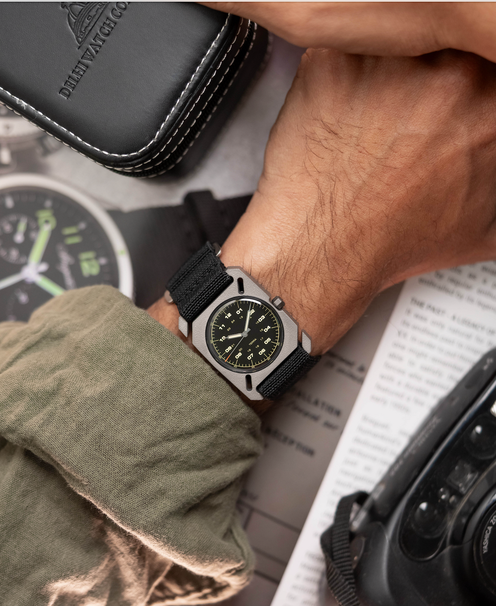 DWC Terra titanium watch with black dial & strap on a wrist. Nearby: Delhi Watch Co. case, olive fabric, camera lens.