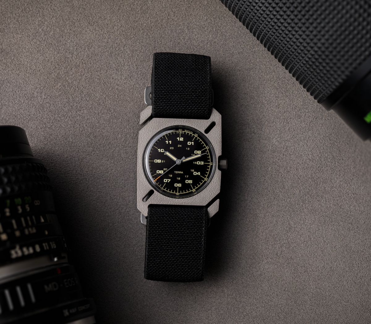 DWC Terra watch: gray titanium square case, black dial with yellow markers, black textile strap. On gray surface with camera lenses.