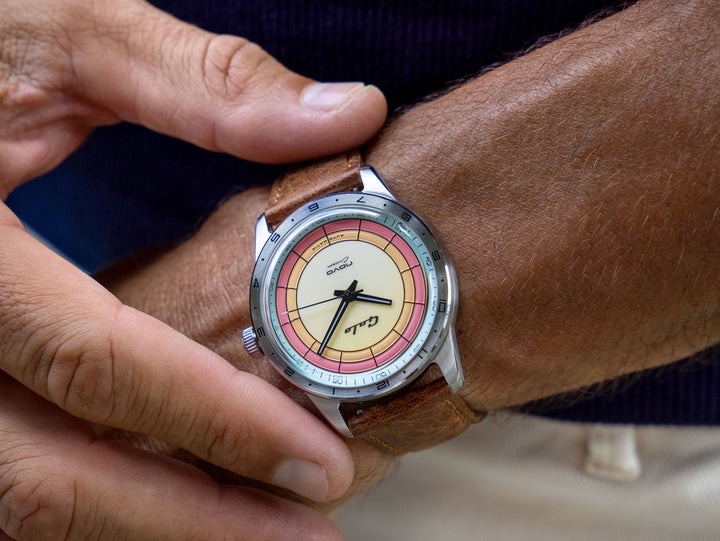 Close-up: Hand adjusts a Galo Novo Cruisin' watch. It has a silver case, brown leather strap, and a cream, yellow, pink concentric dial.