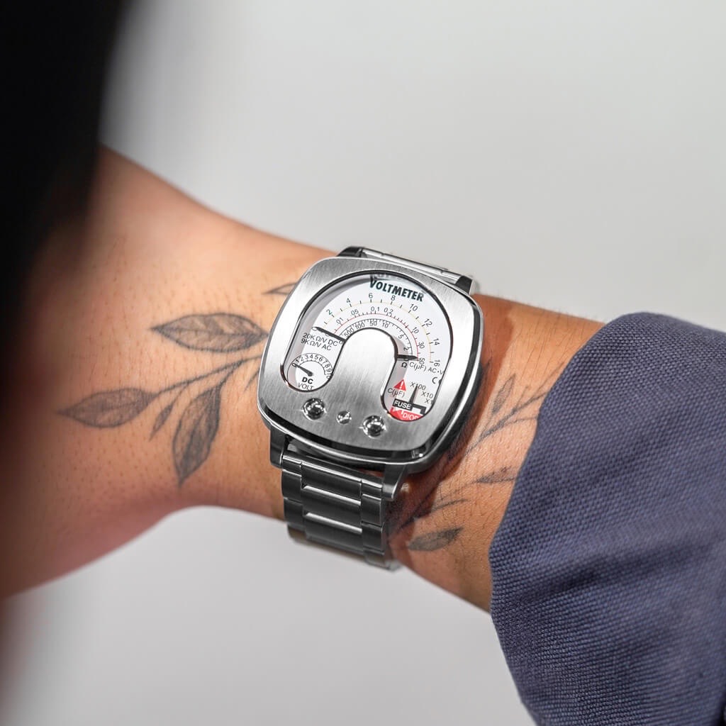 Tattooed arm wears a silver, square watch with a voltmeter-style dial and brushed metal bracelet.