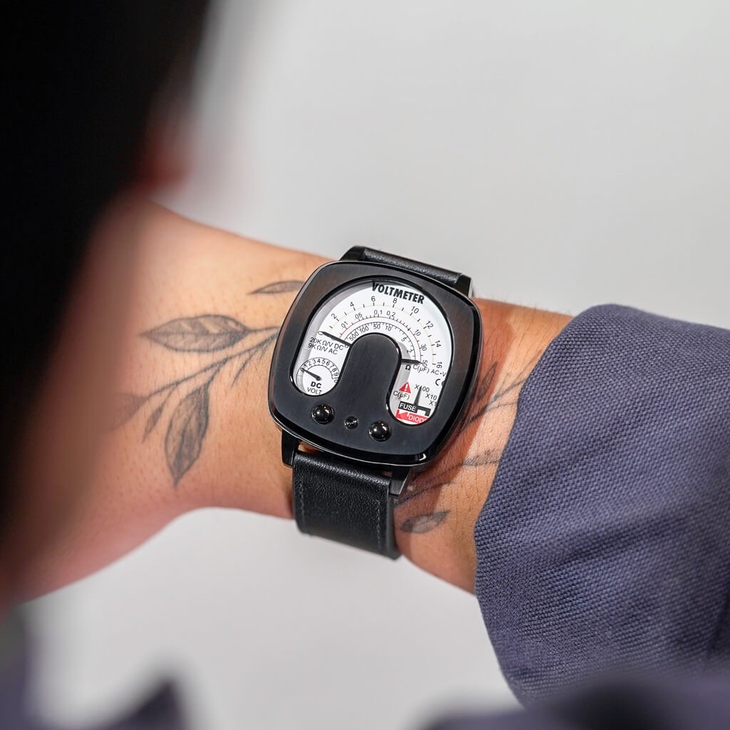 Close-up of a person's wrist wearing a black square Tokyoflash Voltmeter watch with a white analog dial. A leaf tattoo is on the inner wrist.