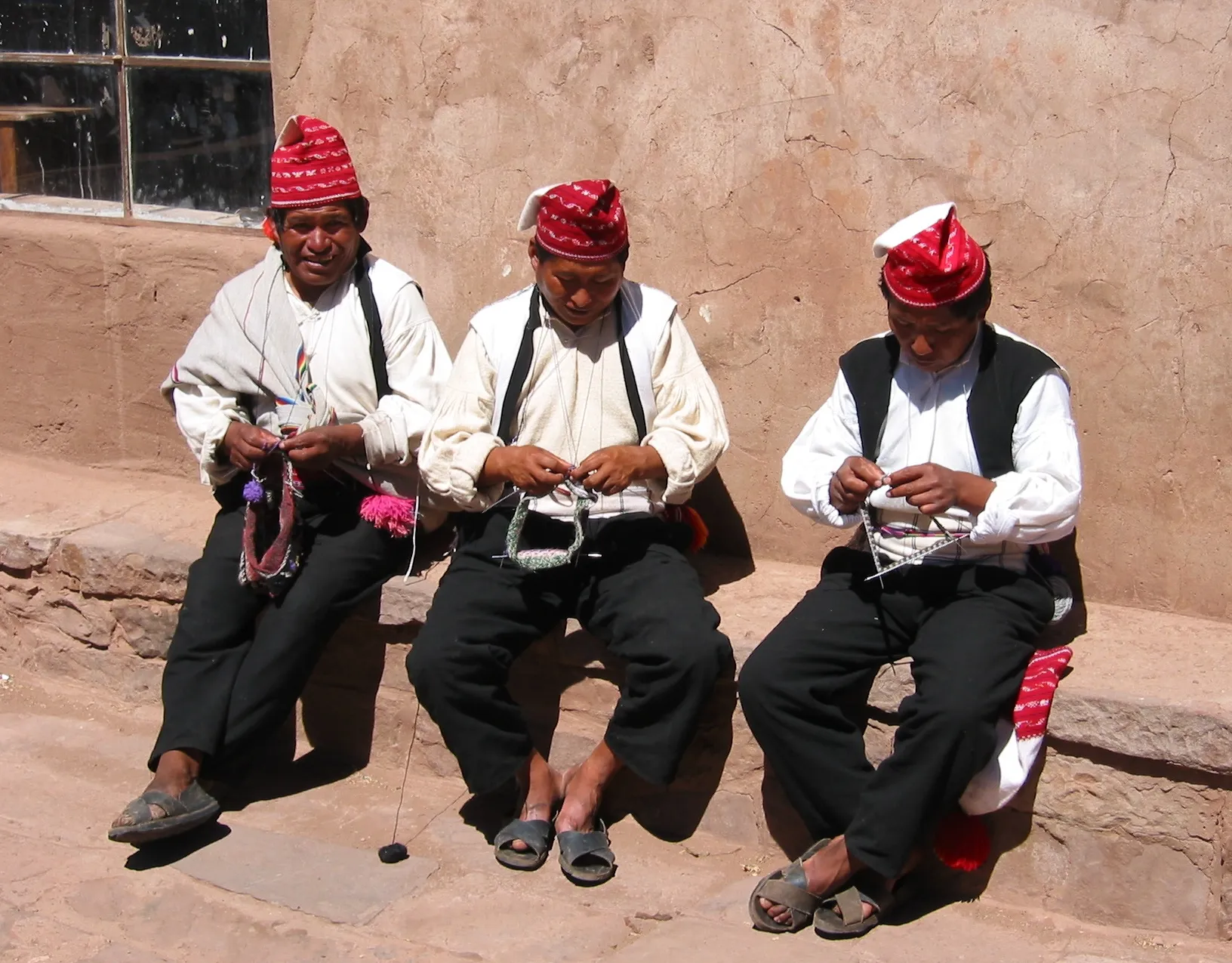 Tres hombres de Taquile sentados en una banca, tejiendo con gorros rojos y camisas blancas