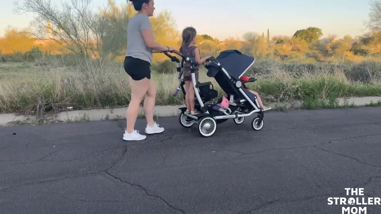 Emily pushing the Austlen Entourage sit-and-stand stroller with an older child standing on the rear platform and a younger child seated in front, on a paved path at sunset.