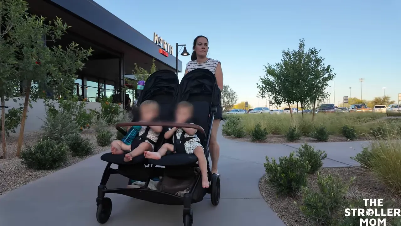 Emily pushing the Bombi Twin side-by-side double stroller with two children seated next to each other, walking along a sidewalk outside a shopping center.