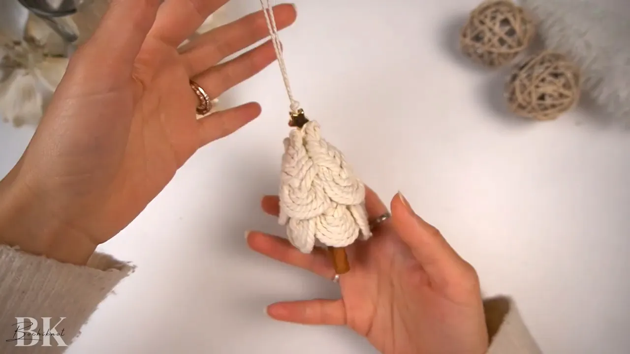 Top-down view of hands holding assembled macrame Pipa knots with a small cinnamon stick in the center while a pink hot glue gun is brought in to apply glue