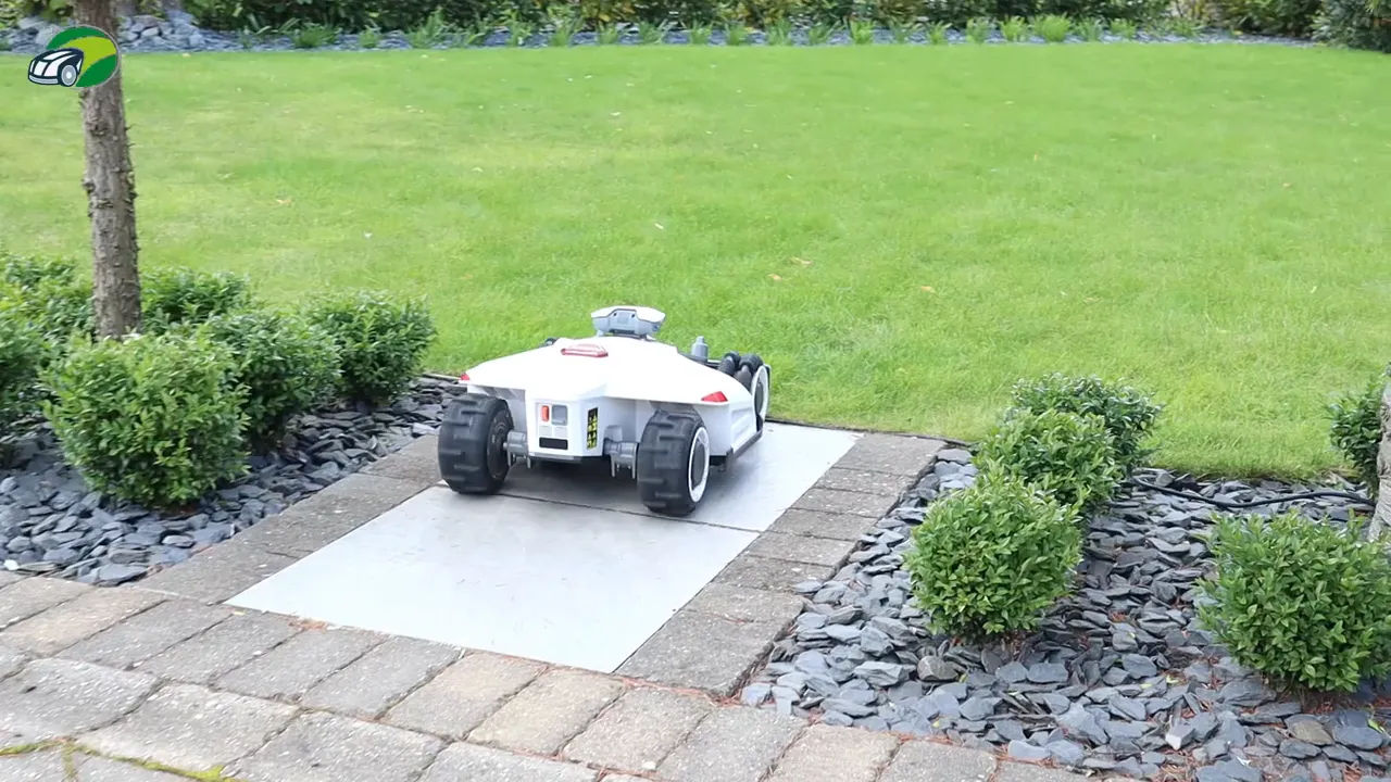 Reviewer using smartphone while Luba 2 robotic mower sits at charging base beside a garden shed, showing docking and alignment