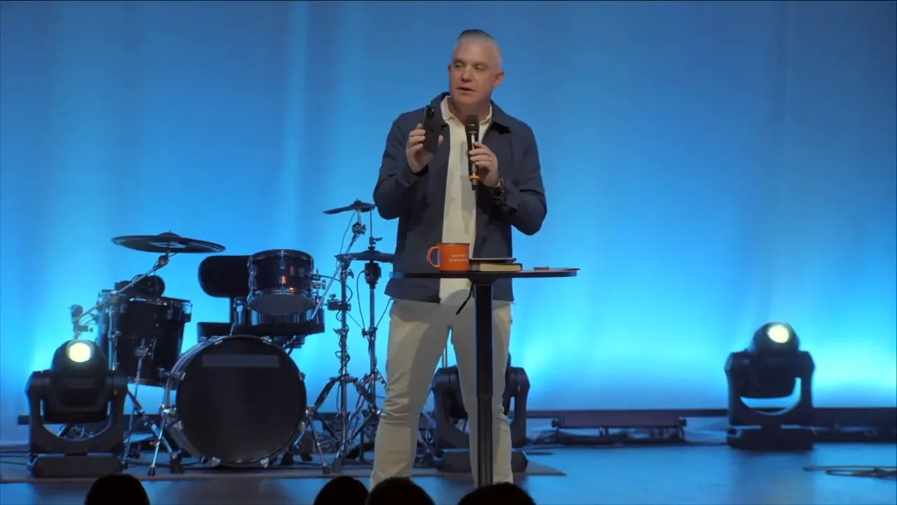 Wide shot of the speaker on stage with 'THE PLACE OF PROVISION' banner and band instruments behind him