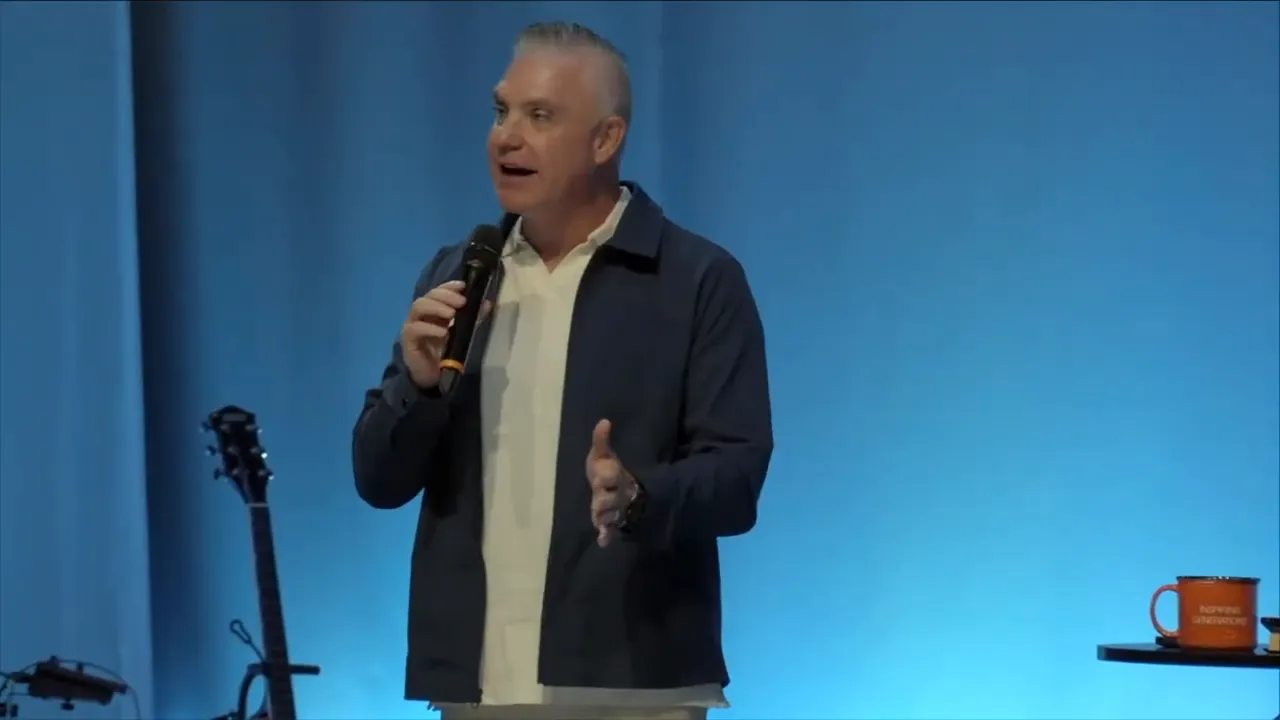 Speaker walking across the stage with a microphone, table with mug and drum kit visible behind.