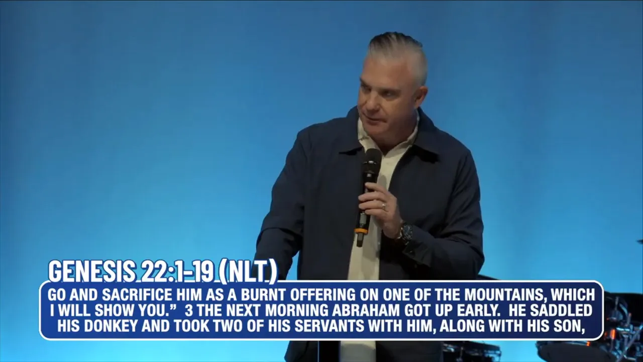 Wide shot of the speaker on stage with 'THE PLACE OF PROVISION' banner and band instruments behind him