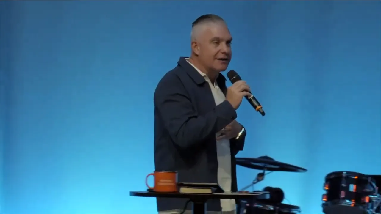 Wide shot of the pastor gesturing with his arm outstretched on stage, band and drummer behind him and audience silhouettes in the foreground.