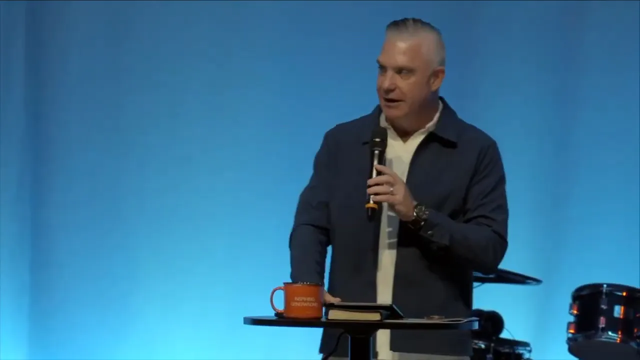 Wide shot of the pastor gesturing with his arm outstretched on stage, band and drummer behind him and audience silhouettes in the foreground.