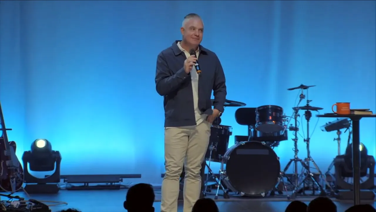 Wide shot of the speaker on stage with 'THE PLACE OF PROVISION' banner and band instruments behind him
