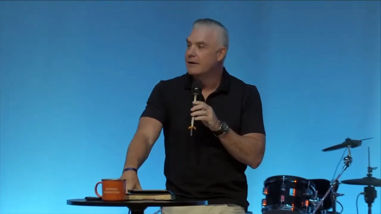 Wide shot of a speaker on stage with blue curtains, audience silhouettes in foreground and a large 'NEXT' graphic on the left screen.