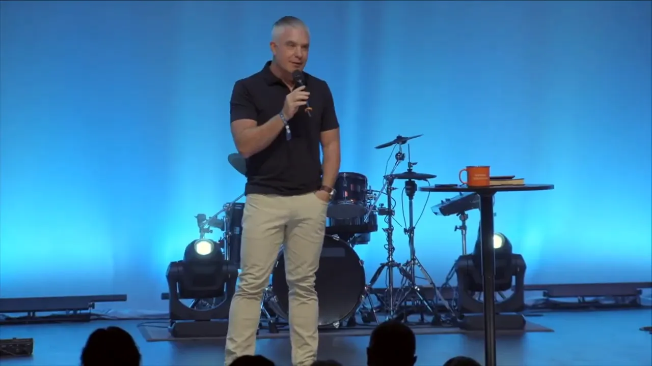 Wide shot of a speaker on stage with blue curtains, audience silhouettes in foreground and a large 'NEXT' graphic on the left screen.