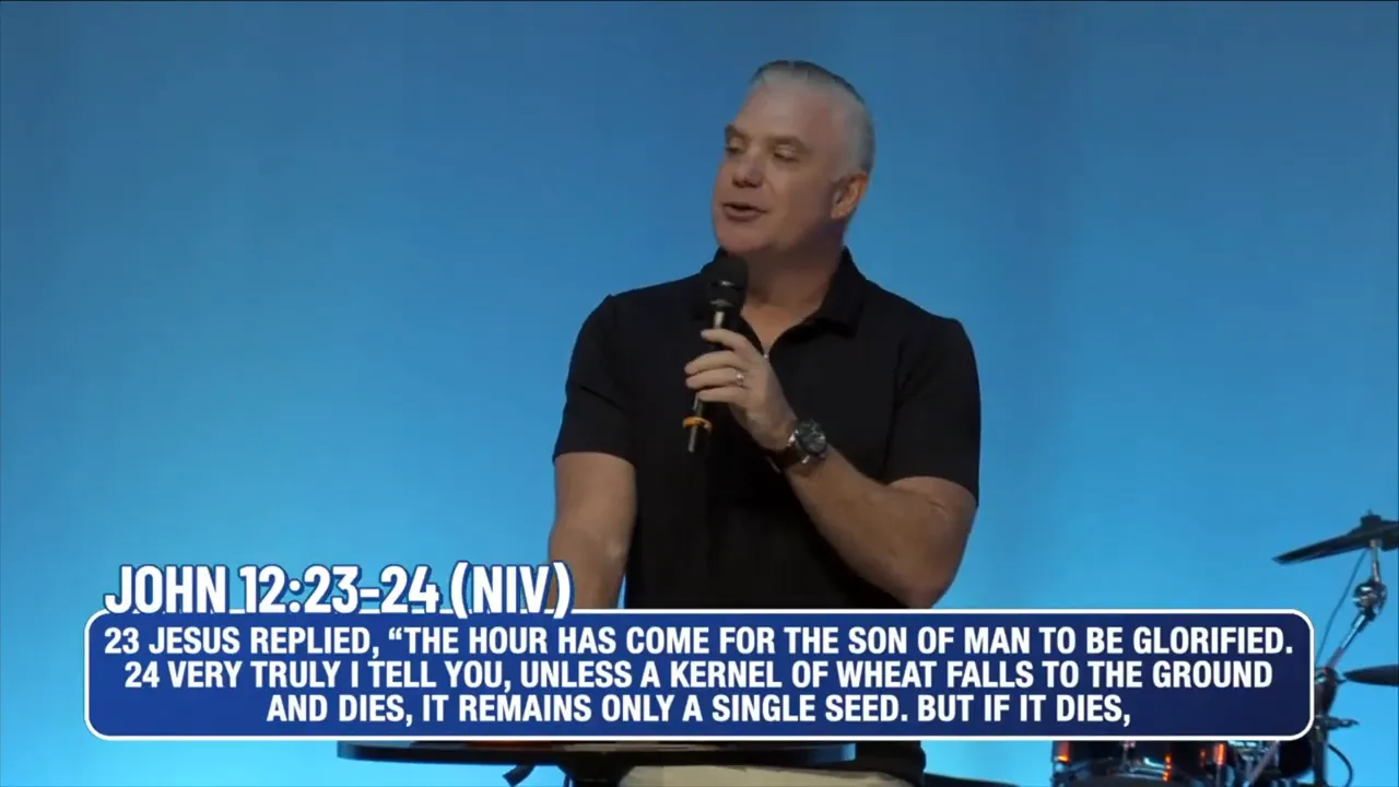 Wide shot of a speaker on stage with blue curtains, audience silhouettes in foreground and a large 'NEXT' graphic on the left screen.