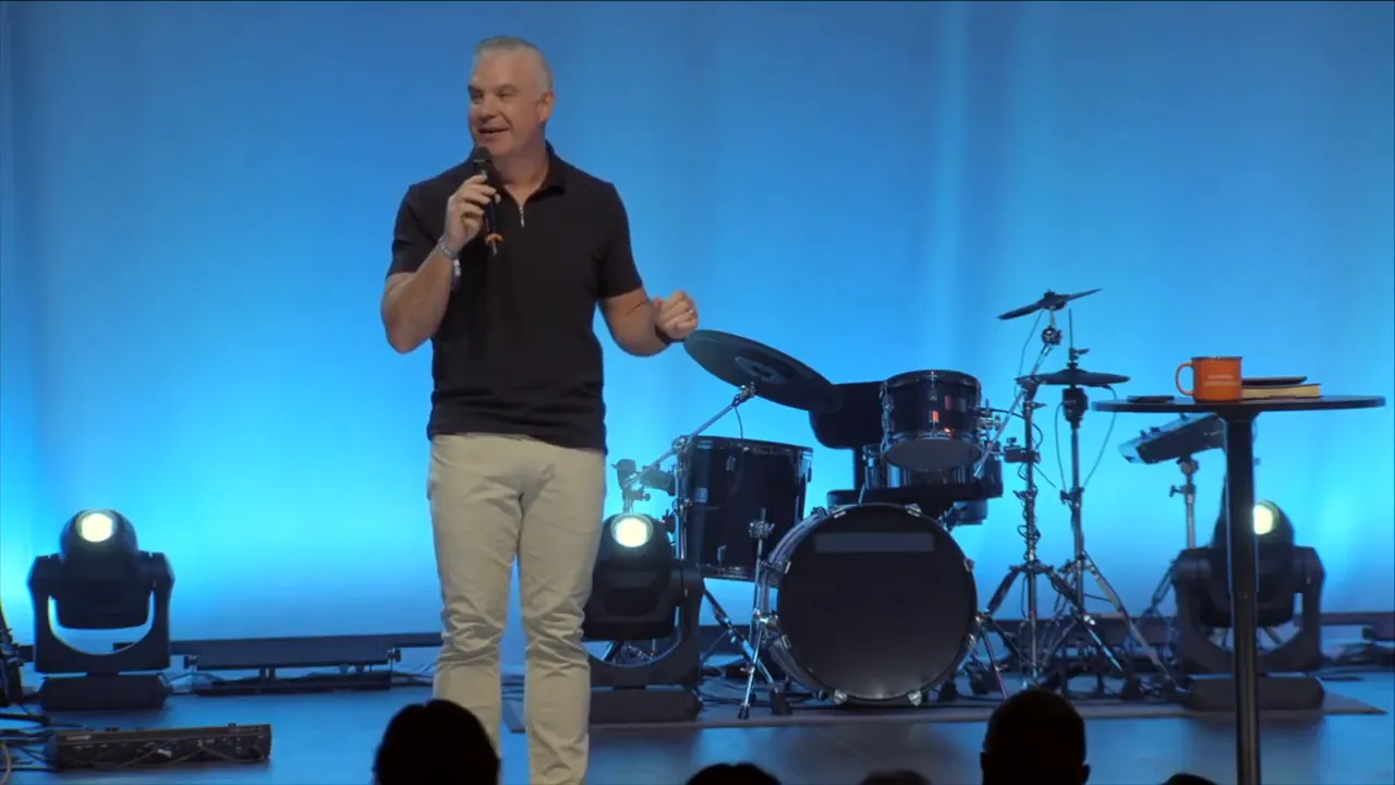 Wide shot of a speaker on stage with blue curtains, audience silhouettes in foreground and a large 'NEXT' graphic on the left screen.