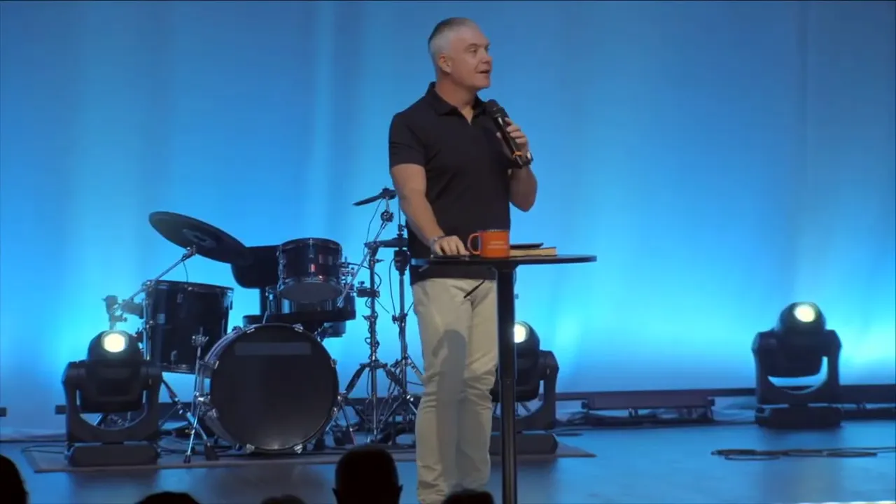 Wide shot of a speaker on stage with blue curtains, audience silhouettes in foreground and a large 'NEXT' graphic on the left screen.