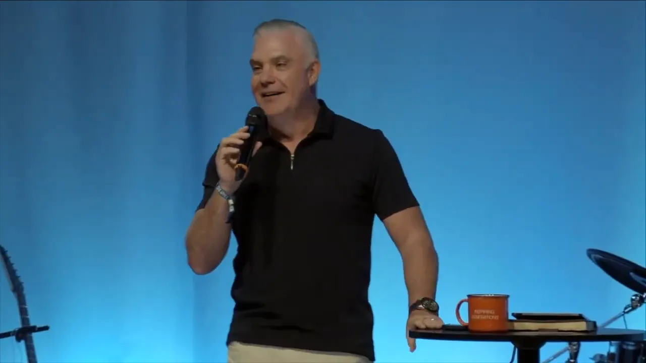 Wide shot of a speaker on stage with blue curtains, audience silhouettes in foreground and a large 'NEXT' graphic on the left screen.