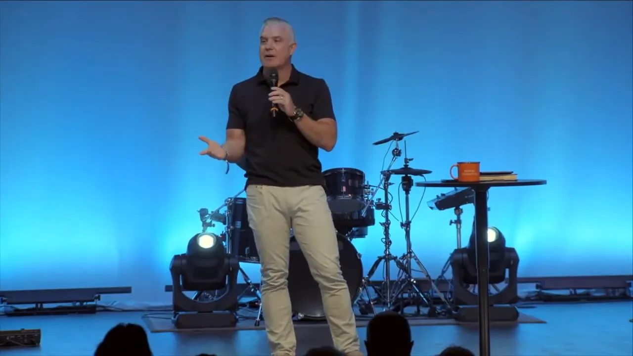 Full stage wide shot with a speaker centered, audience in shadowed foreground and a large vertical screen showing 'NEXT' at left.