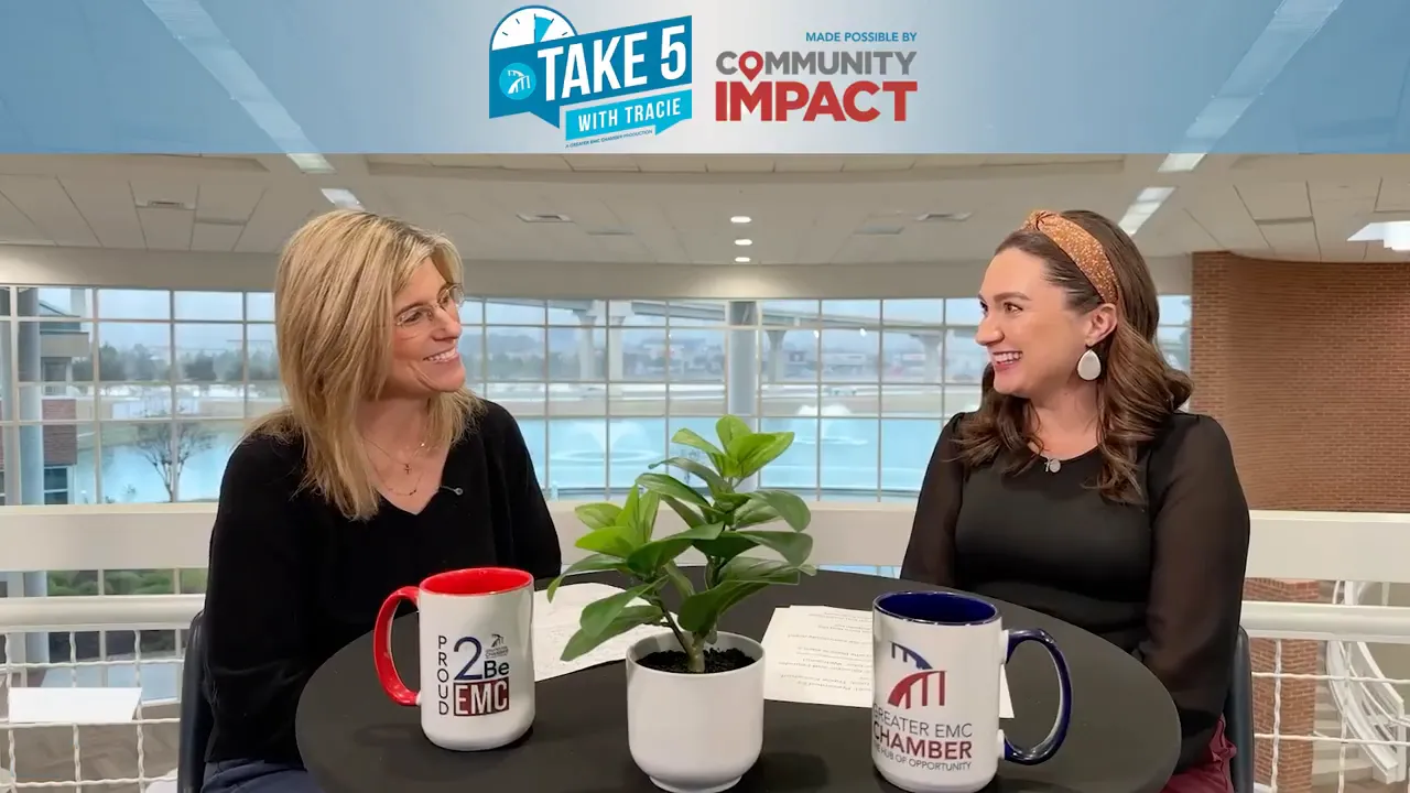 Interview shot of two women at a round table with papers, mugs and a small plant, large windows in the background.