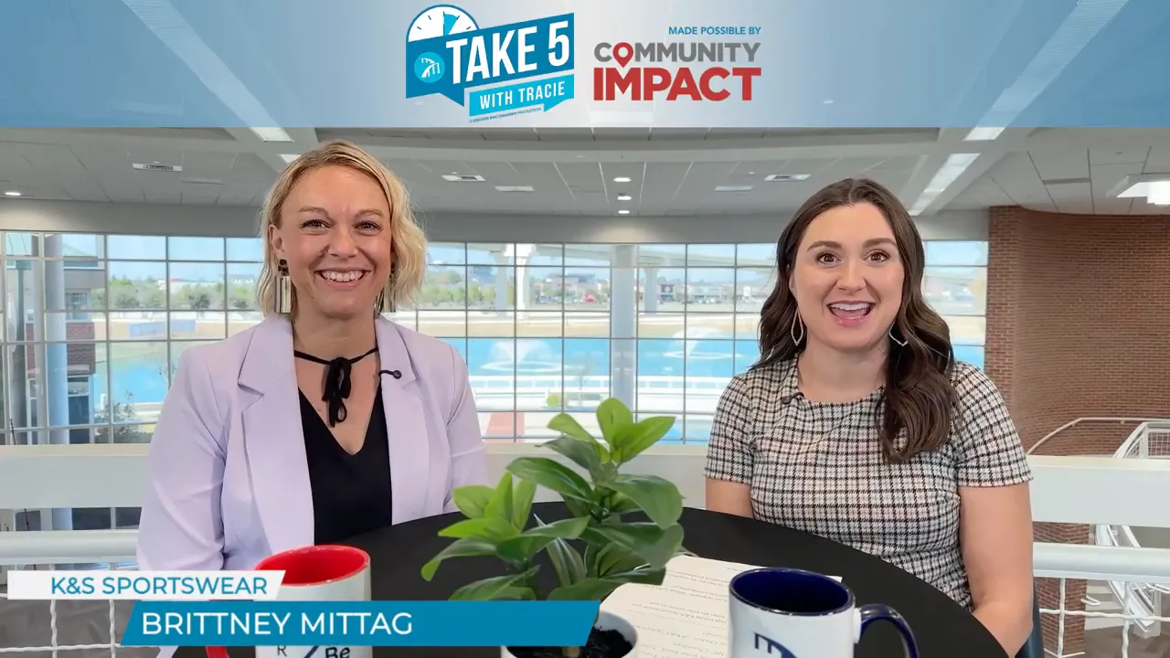 Two women smiling at the camera during a Take 5 interview at a round table with mugs and a plant, bright atrium background.