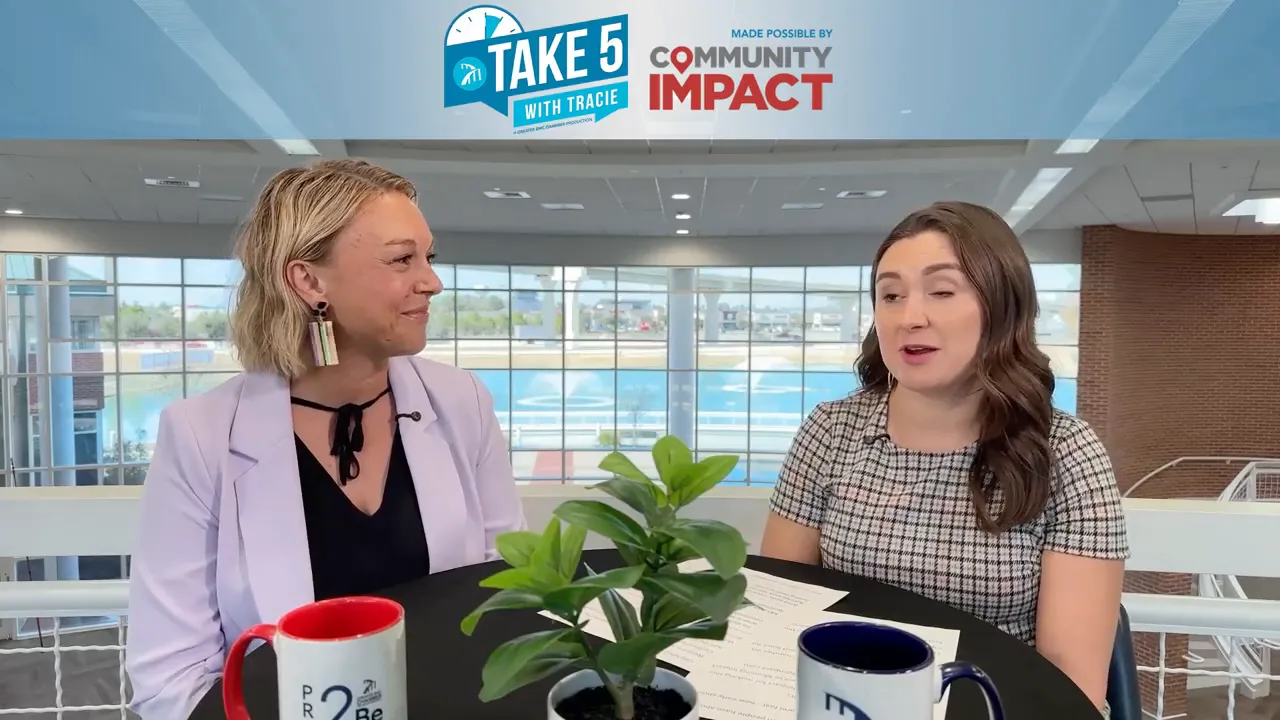 Two people talking at a round table with mugs and a small plant, bright atrium windows behind them