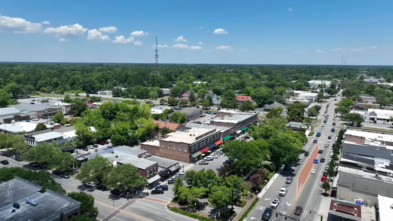 Drone view of three model homes (blue, green, light colored) on a quiet street in the Homecoming community with trees behind.