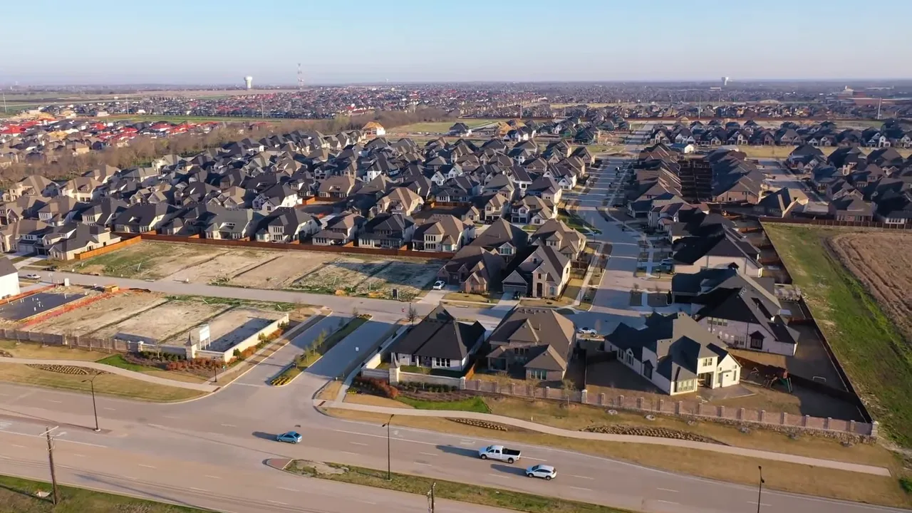Side view of the presenter in a studio with a wood-slat backdrop and a shelf of collectibles, speaking into a desk microphone; clear, well-lit composition showing the recording environment.