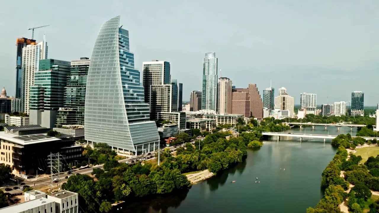 Presenter centered in an outdoor park setting speaking to camera with trees and a building behind him; clear, well-framed shot