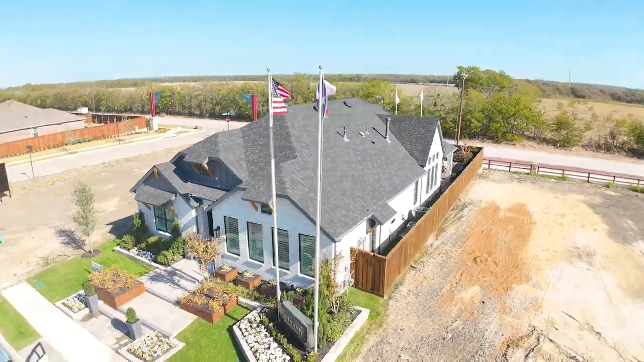 Top-down aerial view of the Highland model home showing roofline, landscaping, flags, and adjacent vacant lots