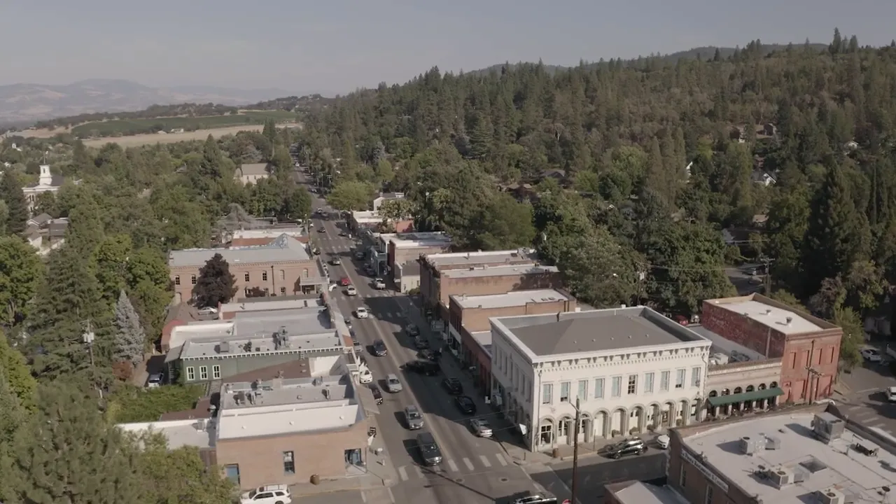 Presenter looking directly at camera with hands together, lightbox reading 'BUYING SOUTHERN OREGON' on shelf behind him