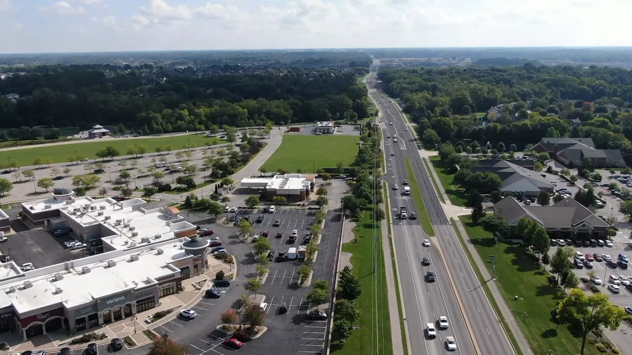 Wide shot of a Noblesville neighborhood edge with open grass, trees, houses and the host speaking